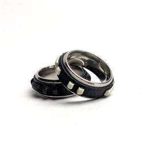 May include: Two stainless steel rings with black bands and silver accents are displayed against a white background. One ring has a textured band, while the other features small, square silver studs. The rings are likely fashion accessories.