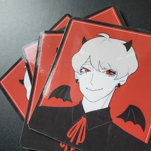 May include: A stack of red and black cards featuring a stylized anime-style character with white hair, red eyes, and small black horns. The character is smiling and has bat wings. The cards are likely part of a game or collectible set.