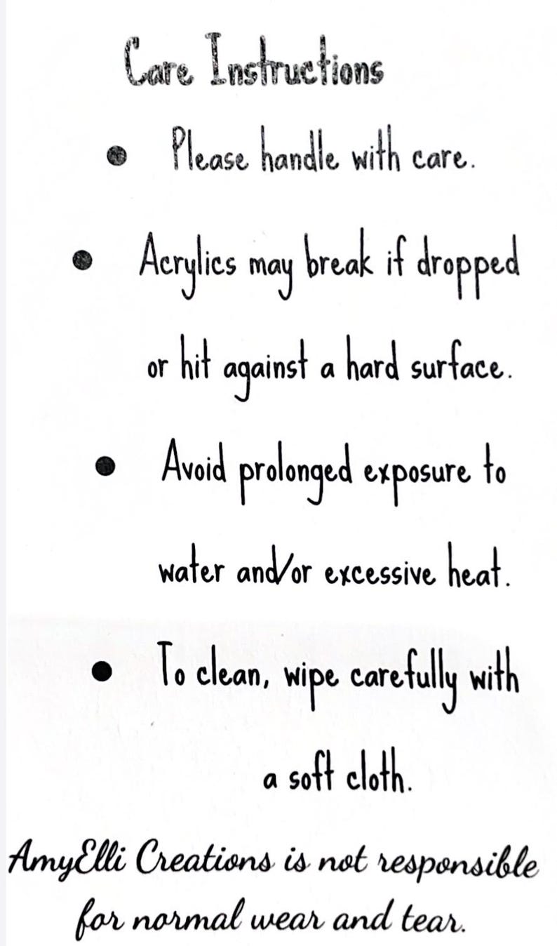 May include: A white background with black handwritten text detailing care instructions. The text advises to handle with care, avoid water and heat, and wipe clean with a soft cloth. It also mentions acrylics may break.