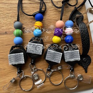 May include: Four retractable badge holders with sports-themed beads. Each holder has a black reel, a silver key ring, and a clear plastic card holder. The beads are in various colours and designs, including baseballs and tennis balls.