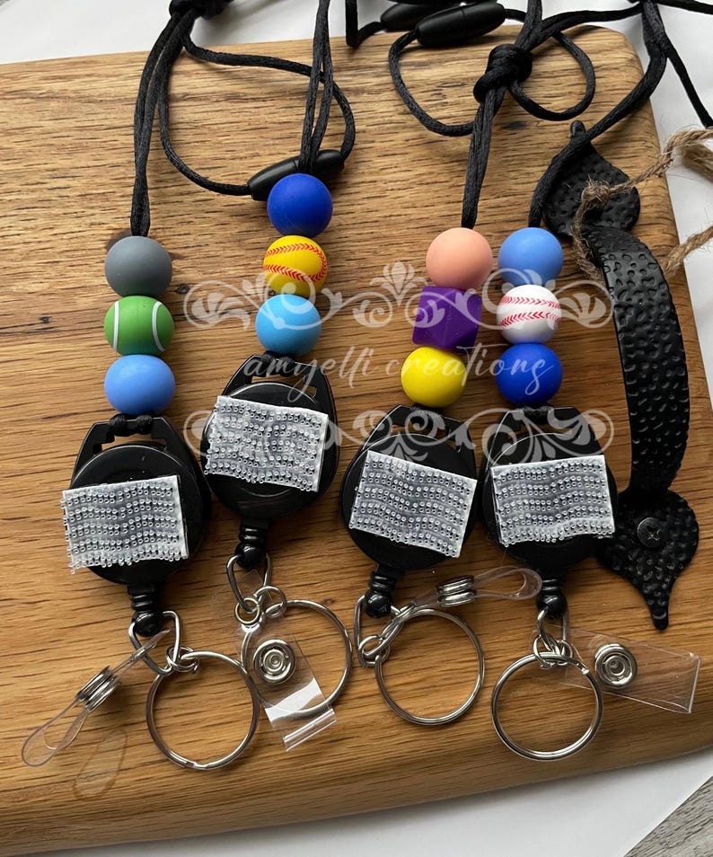 May include: Four retractable badge holders with sports-themed beads. Each holder has a black reel, a silver key ring, and a clear plastic card holder. The beads are in various colours and designs, including baseballs and tennis balls.