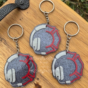 May include: Three keychains with a firefighter theme. Each keychain features a gray, glittery design with a white headset and a red Maltese cross. The keychains have silver-colored key rings and chains. The background is a wooden surface.