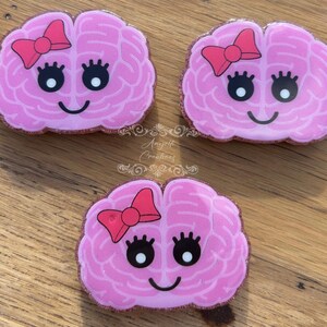 May include: Three pink brain-shaped accessories with a bow, eyelashes, and a smile. The accessories have a glittery border and are arranged on a wooden surface. The accessories are likely pins or magnets.