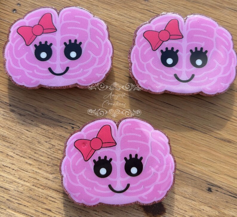 May include: Three pink brain-shaped accessories with a bow, eyelashes, and a smile. The accessories have a glittery border and are arranged on a wooden surface. The accessories are likely pins or magnets.