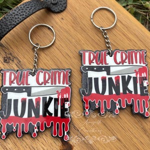 May include: Two keychains with the text "TRUE CRIME JUNKIE" in red and black, featuring a knife graphic and dripping blood design. Each keychain has a silver chain and ring.