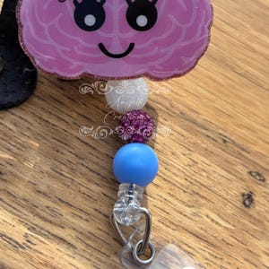 May include: A pink brain-shaped badge reel with a smiling face and a pink bow. The badge reel has a series of beads in white, purple, and blue. The badge reel is attached to a clear plastic clip.