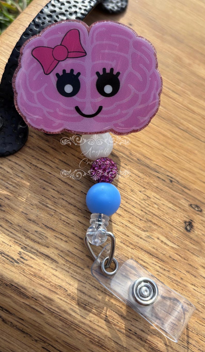 May include: A pink brain-shaped badge reel with a smiling face and a pink bow. The badge reel has a series of beads in white, purple, and blue. The badge reel is attached to a clear plastic clip.