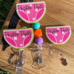 May include: Two pink and green glitter badge holders with the text "Positive Pants" and a pink underwear graphic. The badge holders are attached to clear retractable badge reels with silver clips.