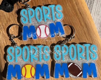 May include: Three glitter-filled keychains with the text "SPORTS MOM" in blue and white lettering. Each keychain features a different sports ball: a baseball, a softball, and a football.