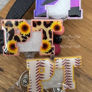 May include: Three badge reels with the letters "PT". One has a purple and black glitter design, another has a leopard print with sunflowers, and the last has a baseball design with red stitching.