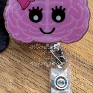 May include: A pink brain-shaped badge reel with a bow, eyelashes, and a smiling face. The badge reel has a glittery border and a clear plastic clip with a metal clasp. The text "Amy's Creations" is visible.