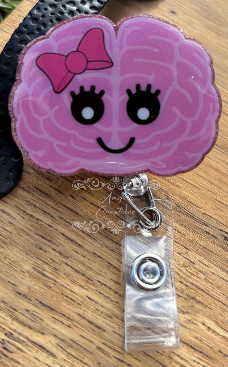 May include: A pink brain-shaped badge reel with a bow, eyelashes, and a smiling face. The badge reel has a glittery border and a clear plastic clip with a metal clasp. The text "Amy's Creations" is visible.