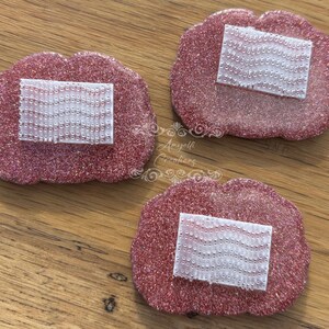 May include: Three pink, glitter-covered, lip-shaped coasters, each with a rectangular, white, beaded detail. They are displayed on a wooden surface. The image is a top-down view.