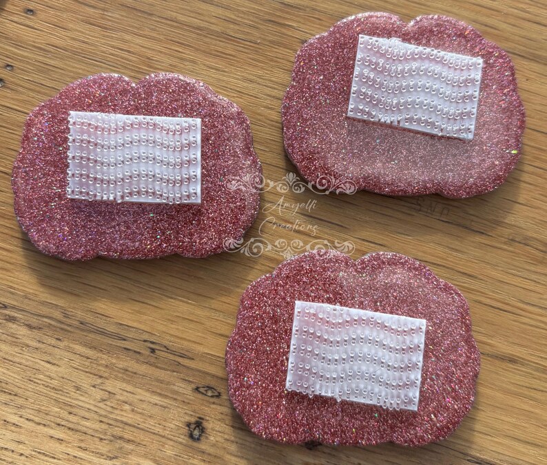 May include: Three pink, glitter-covered, lip-shaped coasters, each with a rectangular, white, beaded detail. They are displayed on a wooden surface. The image is a top-down view.