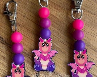 May include: Three keychains with a cartoon dragon design. Each keychain features a silver clasp, a string of pink, purple, and magenta beads, and a dragon charm. The dragon is pink and purple with wings and a friendly expression.