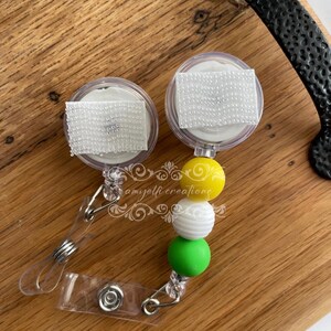 May include: Two clear retractable badge reels with white hook-and-loop fasteners. Each reel has a beaded accent in yellow, white, and green. The reels are on a wooden surface.