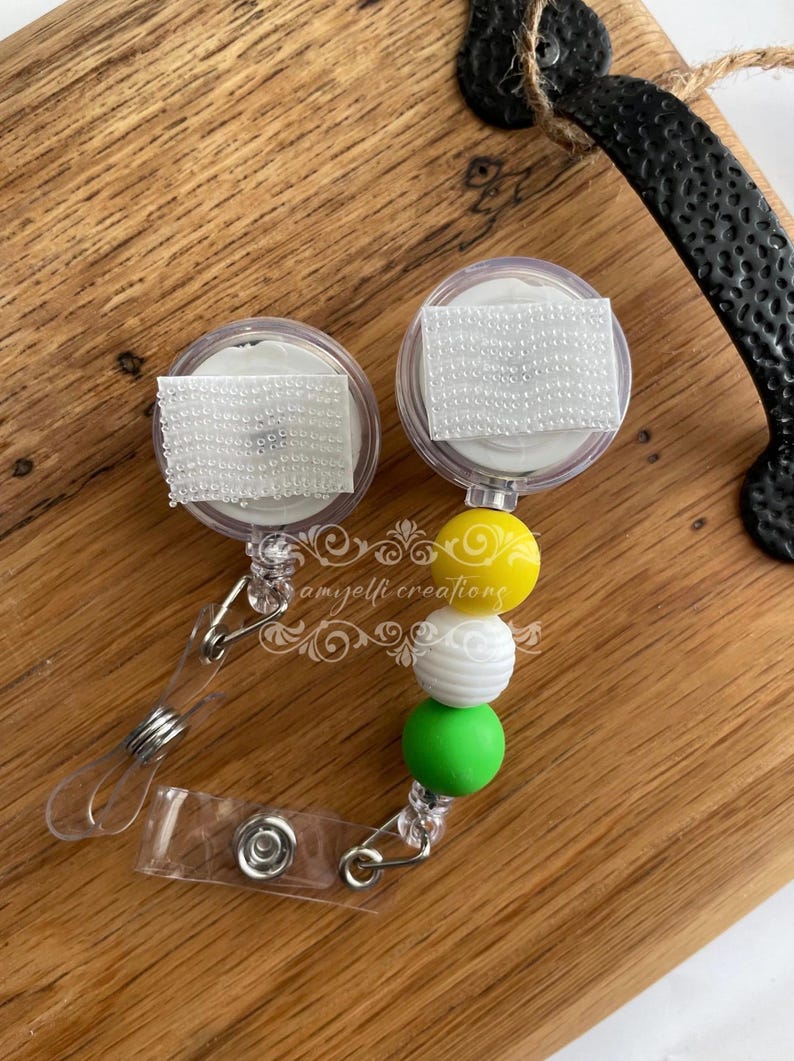 May include: Two clear retractable badge reels with white hook-and-loop fasteners. Each reel has a beaded accent in yellow, white, and green. The reels are on a wooden surface.