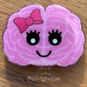 May include: A pink brain-shaped badge with a smiling face, eyelashes, and a bow. The badge has a glittery border and the text "Amyelli Creations" at the bottom. The design is cartoonish and playful.