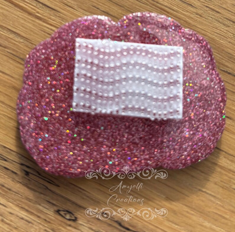 May include: A pink, glitter-covered, lip-shaped tray with a rectangular white object on top. The white object has a dotted pattern. The tray is on a wooden surface. The tray is a handmade item.