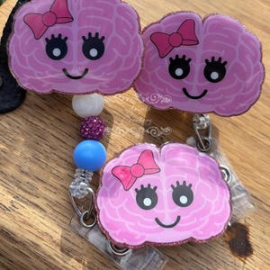 May include: Three pink brain-shaped badge reels with smiling faces and pink bows. The badge reels have a glittery border and are attached to a beaded lanyard with white, purple, and blue beads. The background is a wooden surface.