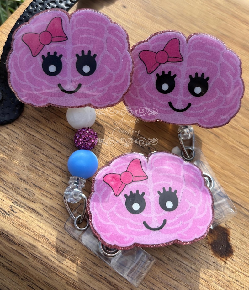 May include: Three pink brain-shaped badge reels with smiling faces and pink bows. The badge reels have a glittery border and are attached to a beaded lanyard with white, purple, and blue beads. The background is a wooden surface.