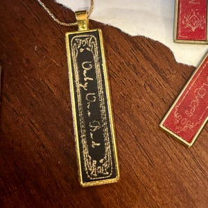 May include: A gold-colored rectangular pendant necklace with the words "Only Us" in gold script on a black background. The pendant is rectangular with decorative gold trim. The necklace is on a wooden surface.