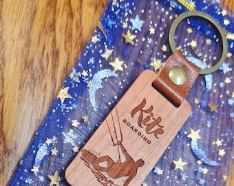 May include: Wooden keychain with a brown leather strap and a silver key ring. The keychain features a silhouette of a person kitesurfing with the text "Kite Boarding" on the wood.