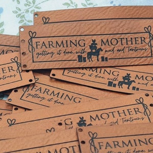 Pu&ograve; includere: Primo piano di diversi cartellini in pelle marrone con il testo "Farming Mother getting it done with mud and tantrums" stampato in nero. I cartellini hanno dei fori punzonati negli angoli e un design decorativo.