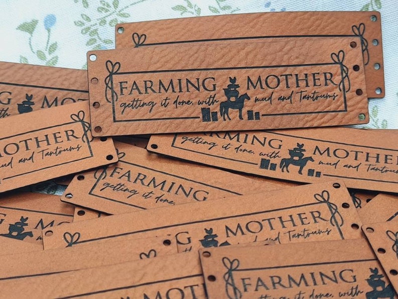Pu&ograve; includere: Primo piano di diversi cartellini in pelle marrone con il testo "Farming Mother getting it done with mud and tantrums" stampato in nero. I cartellini hanno dei fori punzonati negli angoli e un design decorativo.
