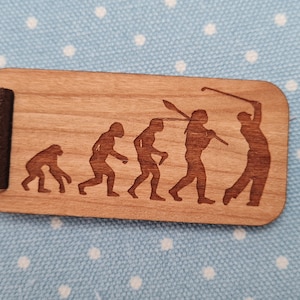 Può includere: Un portachiavi in legno con un'immagine incisa a laser dell'evoluzione dell'uomo, che culmina con un golfista che oscilla una mazza. Il portachiavi ha un cinturino in pelle marrone.