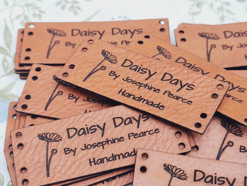 Pu&ograve; includere: Un mucchio di etichette in pelle marrone con il testo "Daisy Days" e "By Josephine Pearce Handmade" stampato su di esse. Ogni etichetta ha un piccolo disegno di margherita.