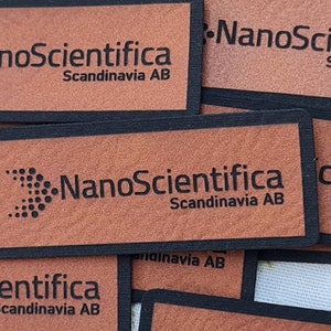 Pu&ograve; includere: Primo piano di quattro etichette in pelle marrone con bordatura nera. Ogni etichetta presenta il testo "NanoScientifica Scandinavia AB" in lettere nere. Le etichette sono impilate l'una sull'altra.