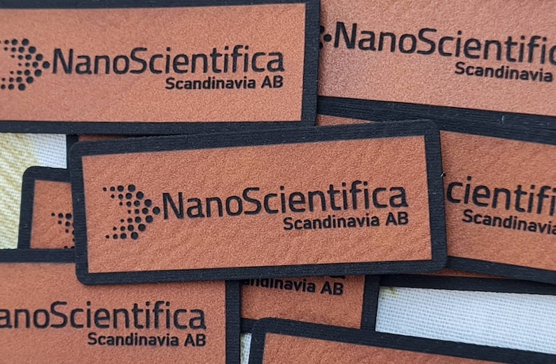 Pu&ograve; includere: Primo piano di quattro etichette in pelle marrone con bordatura nera. Ogni etichetta presenta il testo "NanoScientifica Scandinavia AB" in lettere nere. Le etichette sono impilate l'una sull'altra.