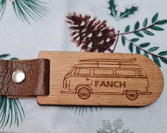Puede incluir: Llavero de madera con correa de cuero marrón y anilla de metal plateado. La etiqueta de madera presenta una imagen grabada de una furgoneta vintage con tablas de surf y el nombre "FANCH".