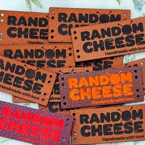 Pu&ograve; includere: Un mucchio di etichette in pelle marrone con il testo "RANDOM CHEESE" in lettere nere e arancioni. Le etichette includono anche il testo "Handmade with love".