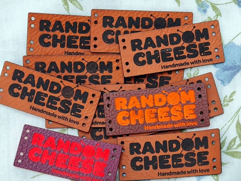 Pu&ograve; includere: Un mucchio di etichette in pelle marrone con il testo "RANDOM CHEESE" in lettere nere e arancioni. Le etichette includono anche il testo "Handmade with love".
