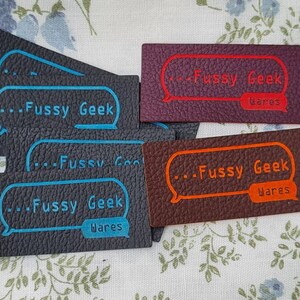 Pu&ograve; includere: Quattro etichette in pelle con il testo "...Fussy Geek Wares" in un fumetto. Le etichette sono di diversi colori: nero, marrone, marrone scuro e viola. Il testo &egrave; in bianco con un contorno blu.