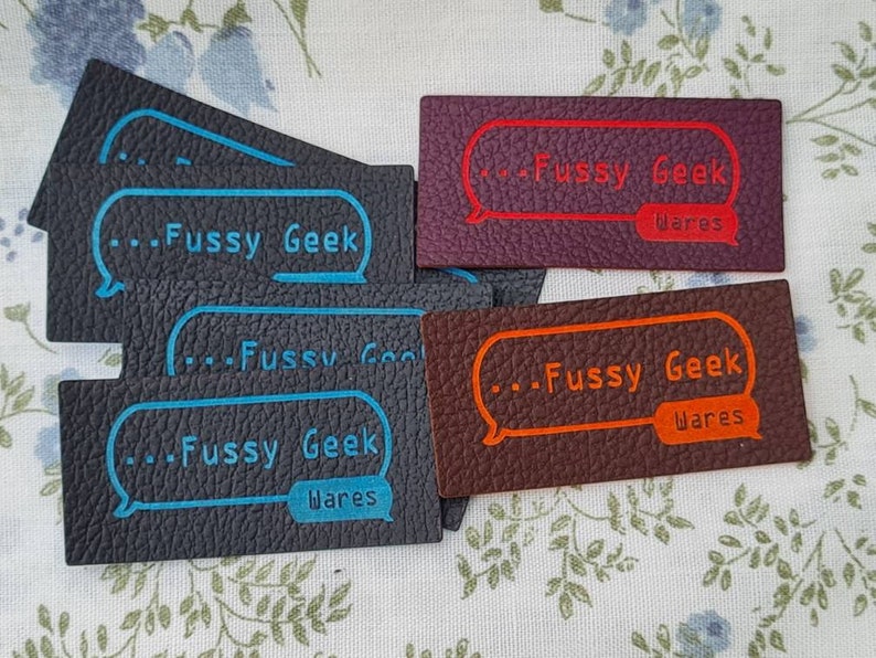 Pu&ograve; includere: Quattro etichette in pelle con il testo "...Fussy Geek Wares" in un fumetto. Le etichette sono di diversi colori: nero, marrone, marrone scuro e viola. Il testo &egrave; in bianco con un contorno blu.