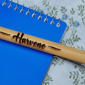 May include: A bamboo pen with a silver tip and a black inscription that reads "Harvene" on a blue notebook.