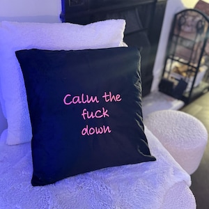 May include: A black cushion with the pink text "Calm the fuck down". It rests on a white, fluffy surface, with a white cushion behind it. A dark wooden headboard is in the background.