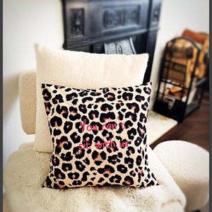 May include: A cushion with a leopard print design and the pink text "You can't sit with us". It rests on a cream-coloured chair with a matching cushion behind it, creating a cosy aesthetic.