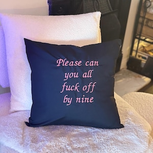 Può includere: Un cuscino nero con la scritta ricamata in rosa "Please can you all fuck off by nine". È appoggiato su una superficie bianca e soffice, con un altro cuscino bianco dietro. Tema: arredamento per la casa.