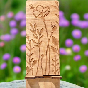 May include: A wooden phone stand with a floral design. The stand features an engraved pattern of flowers, leaves, and bees. The stand is made of light-colored wood and has a rectangular shape with a slot to hold a phone.