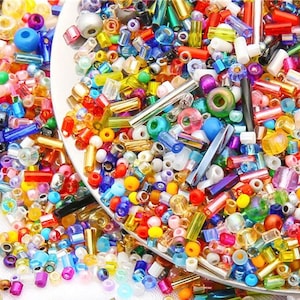 May include: A close-up of a variety of colorful glass beads and seed beads in a bowl. The beads are a mix of shapes and sizes, and they are scattered around the bowl.