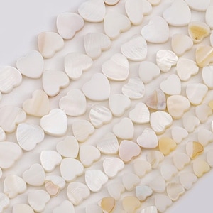 Puede incluir: Una hebra de cuentas de concha en forma de corazón de color blanco y crema. Las cuentas están dispuestas en filas y son de diferentes tamaños.