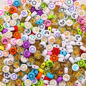 Puede incluir: Una variedad de cuentas de plástico de colores con diferentes formas y diseños, incluyendo estrellas, corazones, lunas y flores. Las cuentas son blancas con contornos negros y algunas tienen detalles de purpurina dorada.