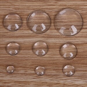 May include: Assortment of ten clear glass cabochons of varying sizes, arranged on a wood surface.