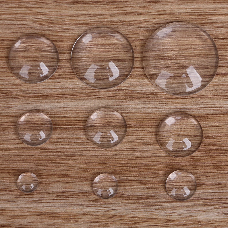 May include: Assortment of ten clear glass cabochons of varying sizes, arranged on a wood surface.