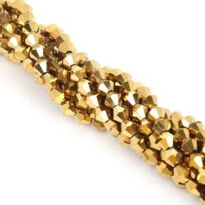 May include: Close-up of a strand of golden, faceted beads. The beads are arranged in a twisted pattern, reflecting light and creating a shimmering effect. The beads are a warm gold color, and the background is white.