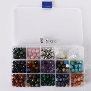 May include: A clear plastic storage box with a blue lid containing a variety of colorful gemstone beads and silver Buddha head charms. The beads are organized in a grid of 12 compartments.